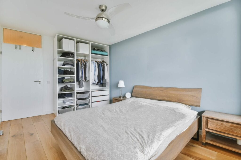Minimalist bedroom featuring a wooden bed, open closet, light blue wall, ceiling fan, and bedside tables—kept spotless with regular home cleaning for a fresh and inviting space.